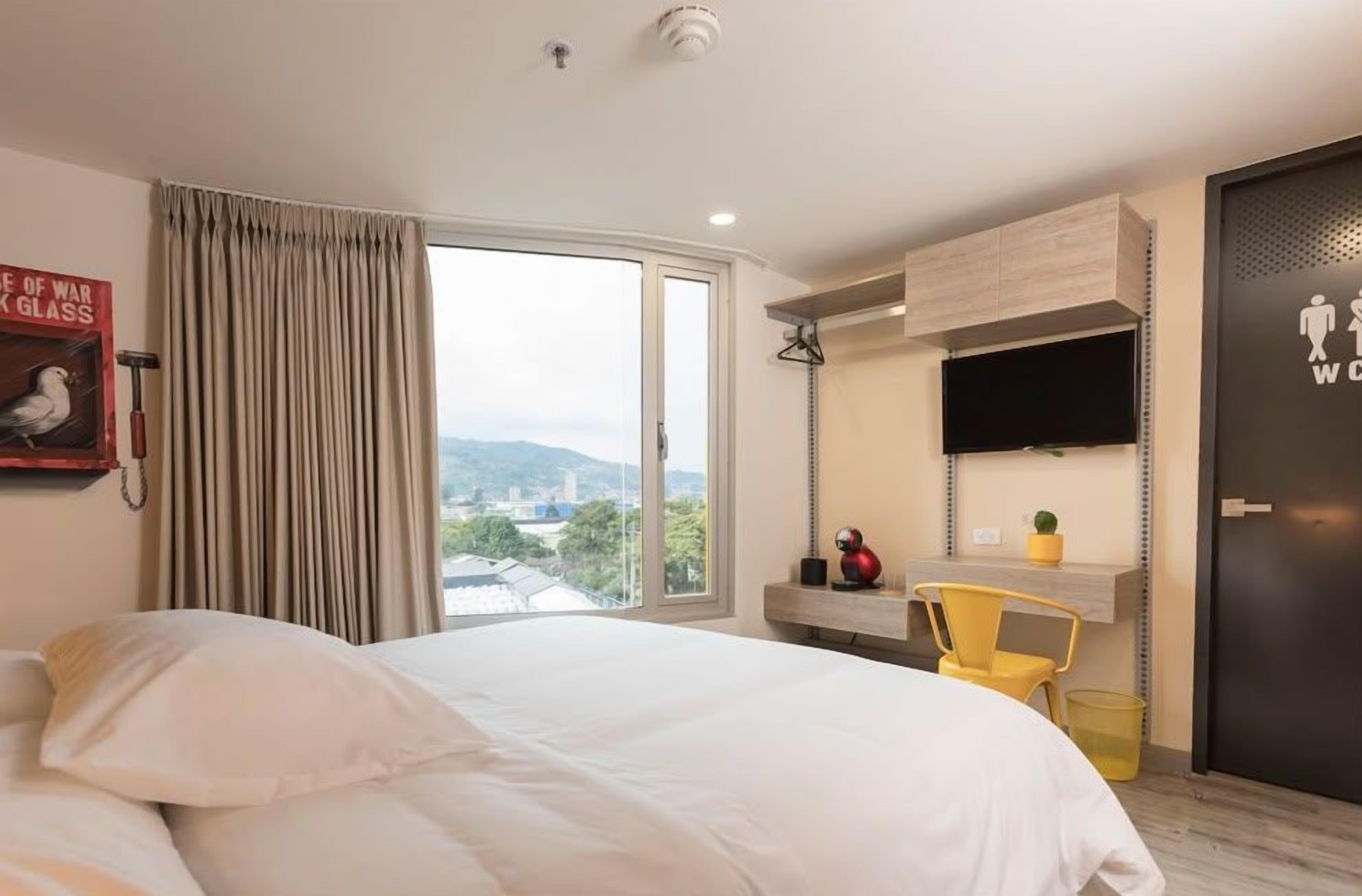 Habitación de hotel con cama, ventana con cortinas, televisor montado, escritorio pequeño con silla y puerta con letrero.