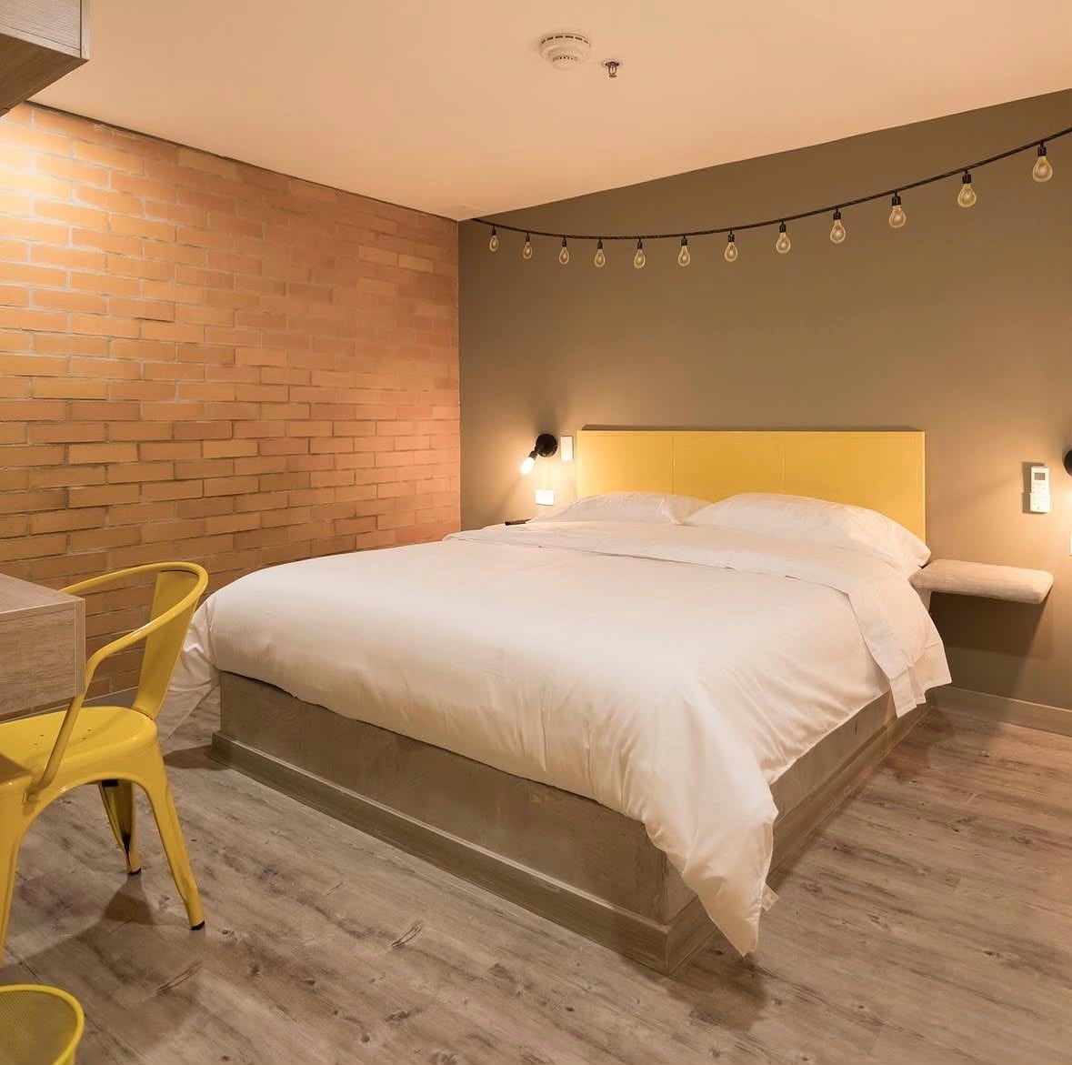 A hotel room with a brick wall, a bed with white bedding, a yellow chair, and a string of lights.