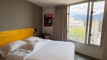 A hotel room with a bed, a window with a view of buildings and mountains, a wall-mounted light, and a fire alarm.