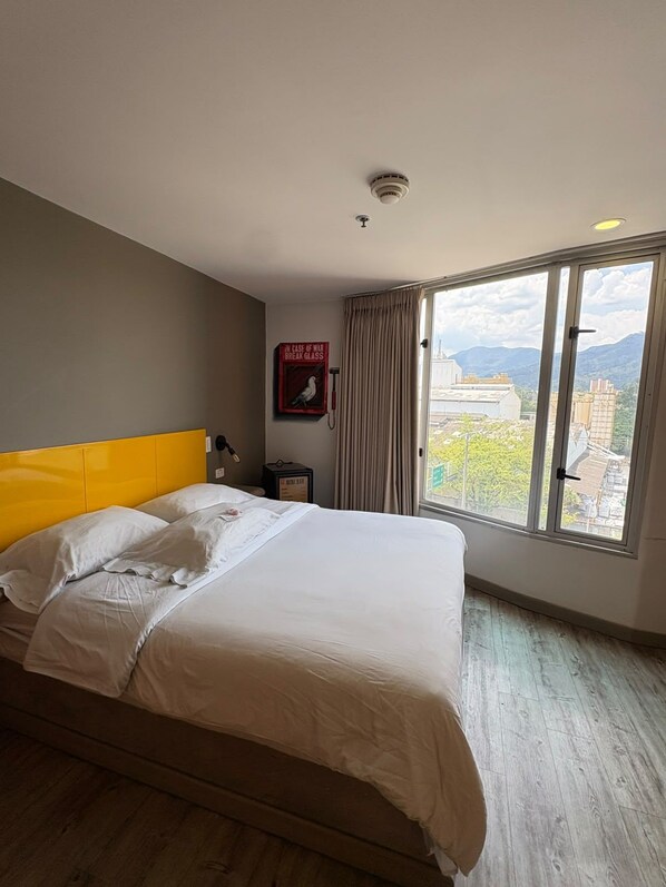 Habitación de hotel con cama, ventana que ofrece vistas a edificios y montañas, lámpara de pared e detector de incendios.