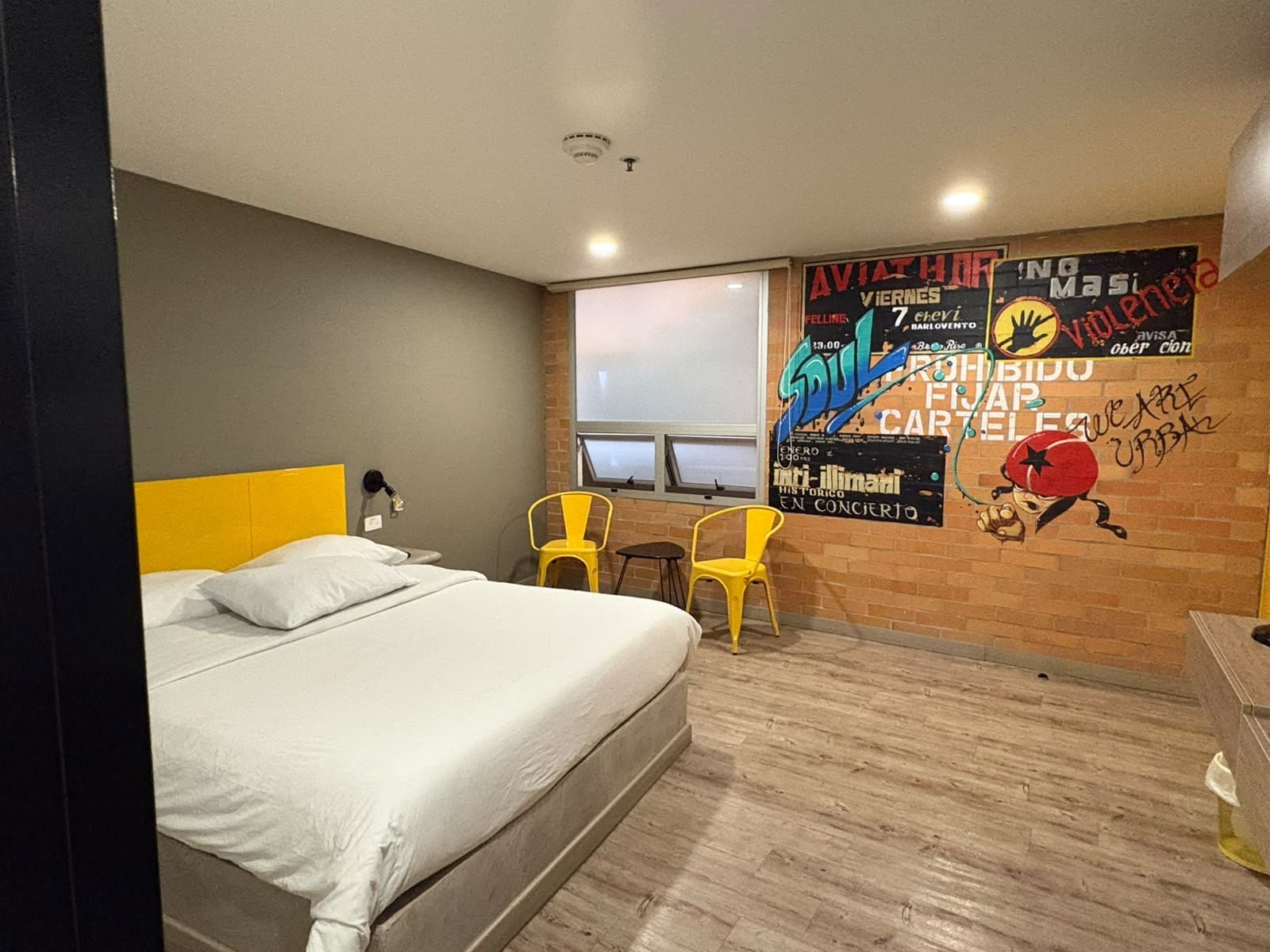 Quarto de hotel com uma cama, duas cadeiras, uma janela e uma parede com um cartaz.