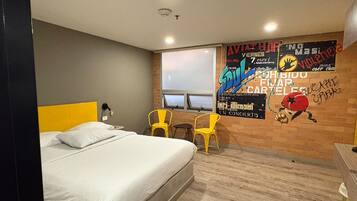 Quarto de hotel com uma cama, duas cadeiras, uma janela e uma parede com um cartaz.