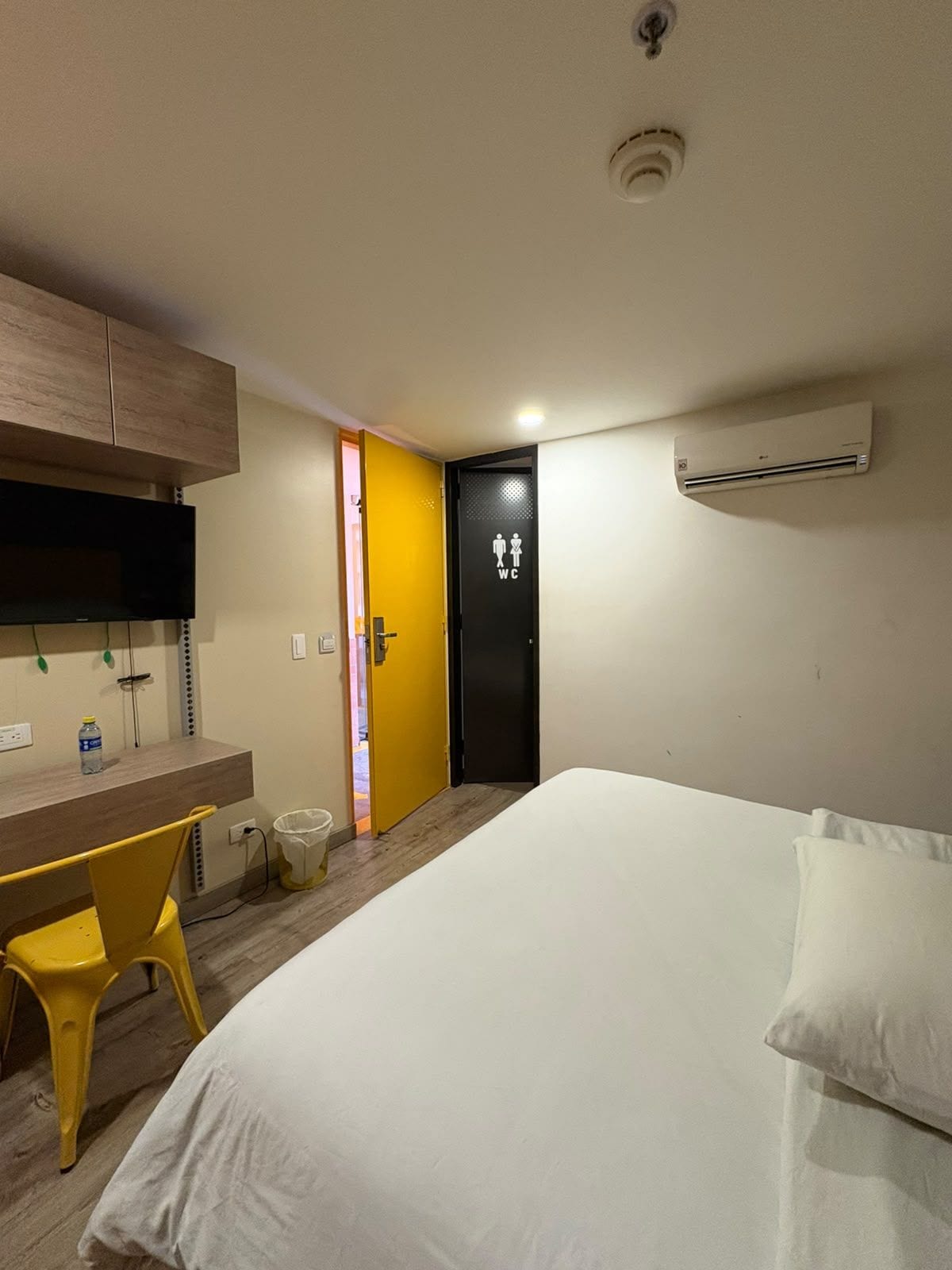 Une chambre d’hôtel équipée d’un lit, d’un bureau, d’une chaise, d’un climatiseur et d’une porte portant le symbole des toilettes.