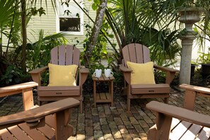 Zwei Adirondack-Stühle mit gelben Kissen, ein kleiner Tisch mit zwei weißen Tassen und ein Steingartenpflanzgefäß in einem Garten.