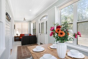 A dining area with a wooden table set for four, a vase of flowers, and a view of the outdoors through large windows. - Walk to Downtown, Shops, Restaurants! Charming Historic Home, Screened Porch, $300 in Free Tickets! (St. Augustine)