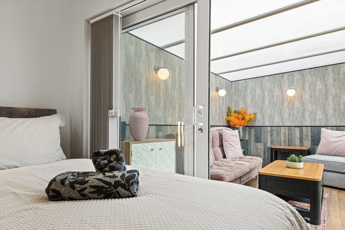 A modern bedroom with a large bed, a glass door leading to a balcony, and a seating area with a sofa and a small table.
