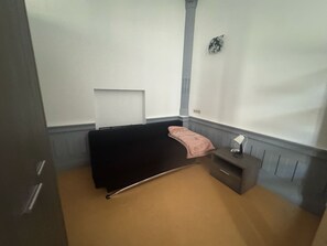 A small, single-bed room with a dark-colored bed, a small bedside table with a lamp, and a plain white wall. - Bright 1-bedroom apartment in Outremeuse, self check-in, near city centre (Liège)