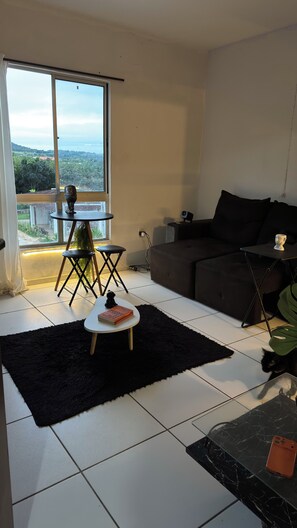A modern living room with a large window, a black sofa, a small round table, and a black rug. - Room with a view of the pool in a secure residential complex (Vitória de Santo Antão)