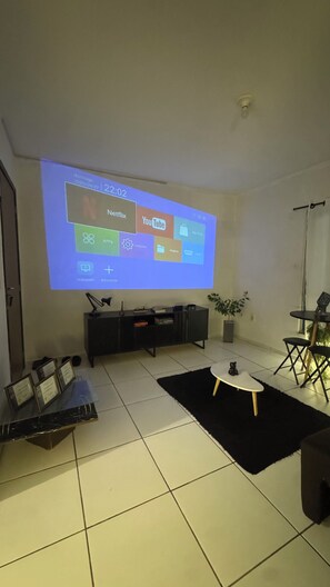 Una sala de estar con una pantalla grande mostrando una interfaz digital, un mueble de entretenimiento negro, una mesita con una planta y una alfombra negra en el suelo.