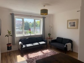 Ett vardagsrum med en soffa, ett litet bord, en krukväxt och ett fönster med gardiner.