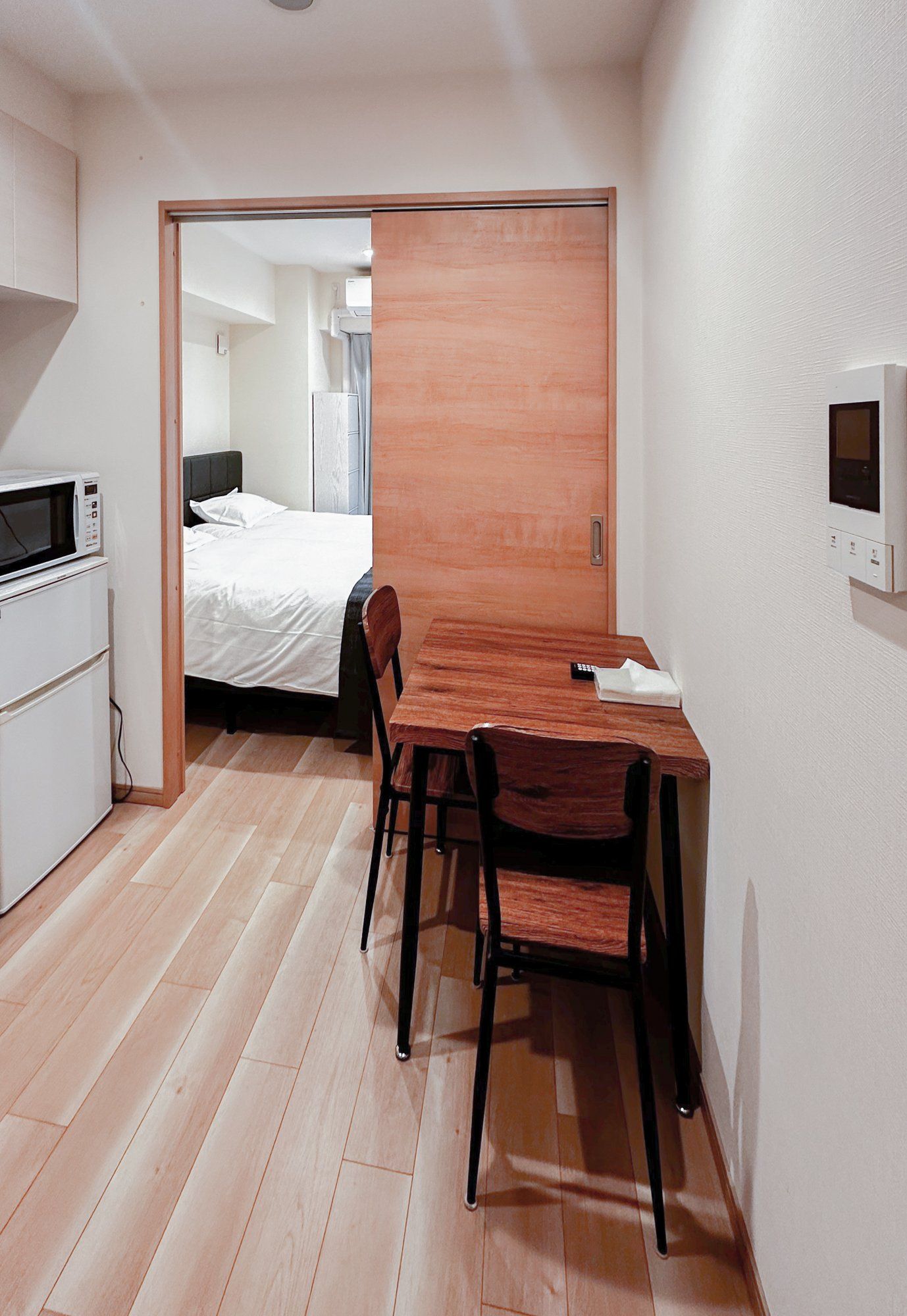 Habitación compacta con una mesa de madera, dos sillas, microondas y una cama con cubrecama blanco.