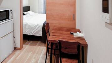 Habitación compacta con una mesa de madera, dos sillas, microondas y una cama con cubrecama blanco.