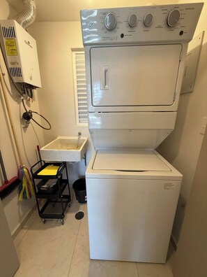 A compact laundry room with a stacked washer and dryer, a small sink, and a cart with cleaning supplies. - Departamento de 1 recámara frente al Estadio TSM, alberca y gimnasio (Torreón)
