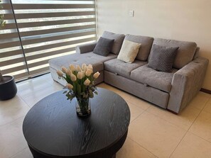 Un salon avec un canapé gris, une table ronde noire ornée d’un vase de fleurs blanches et une plante en pot.