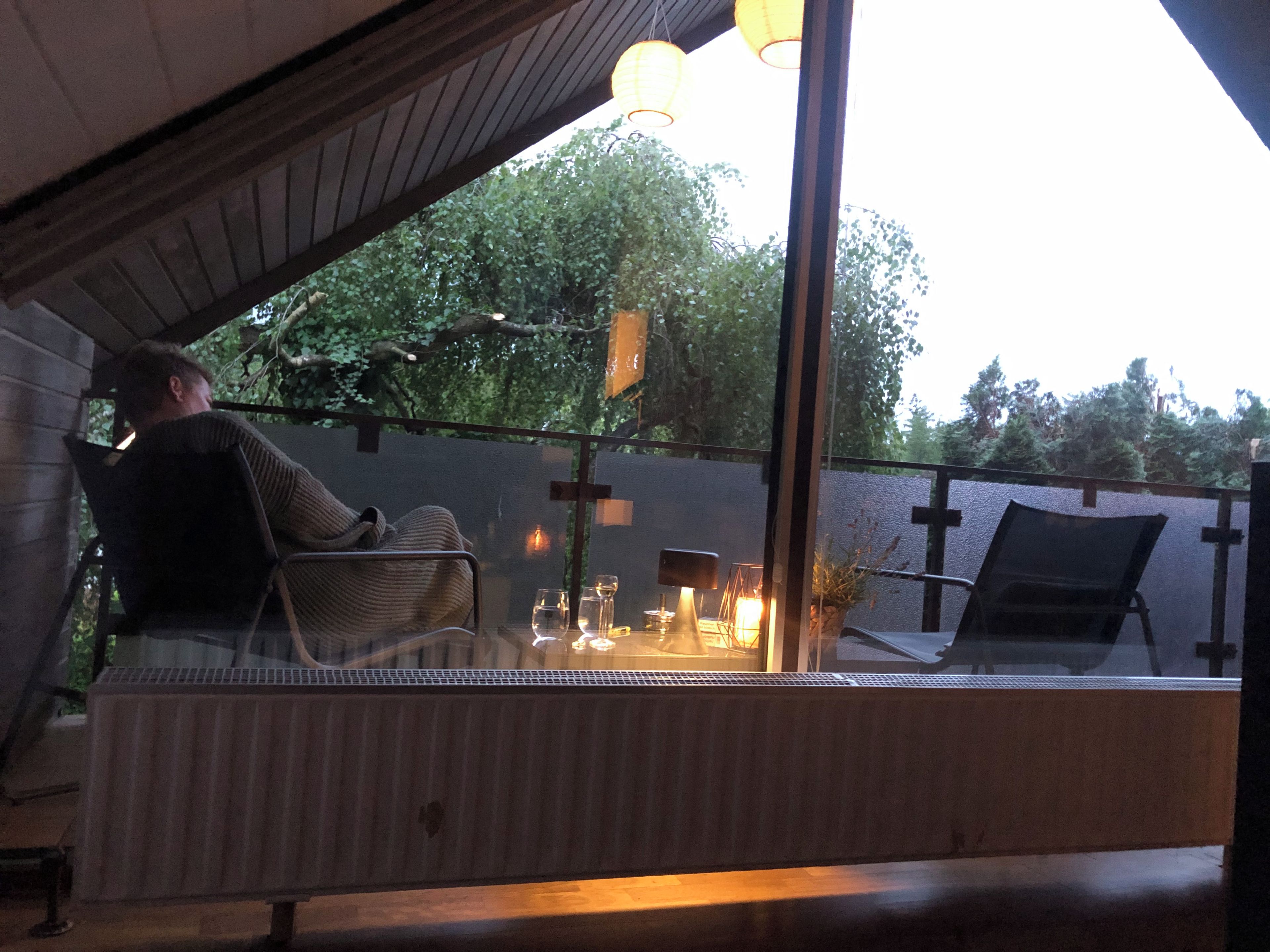 A person seated at a table with a glass of wine, a candle, and a lamp, in a room with a view of trees.