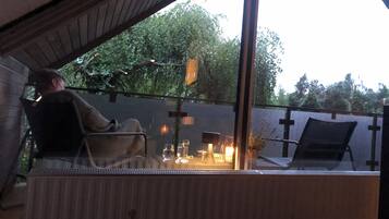 A person seated at a table with a glass of wine, a candle, and a lamp, in a room with a view of trees.