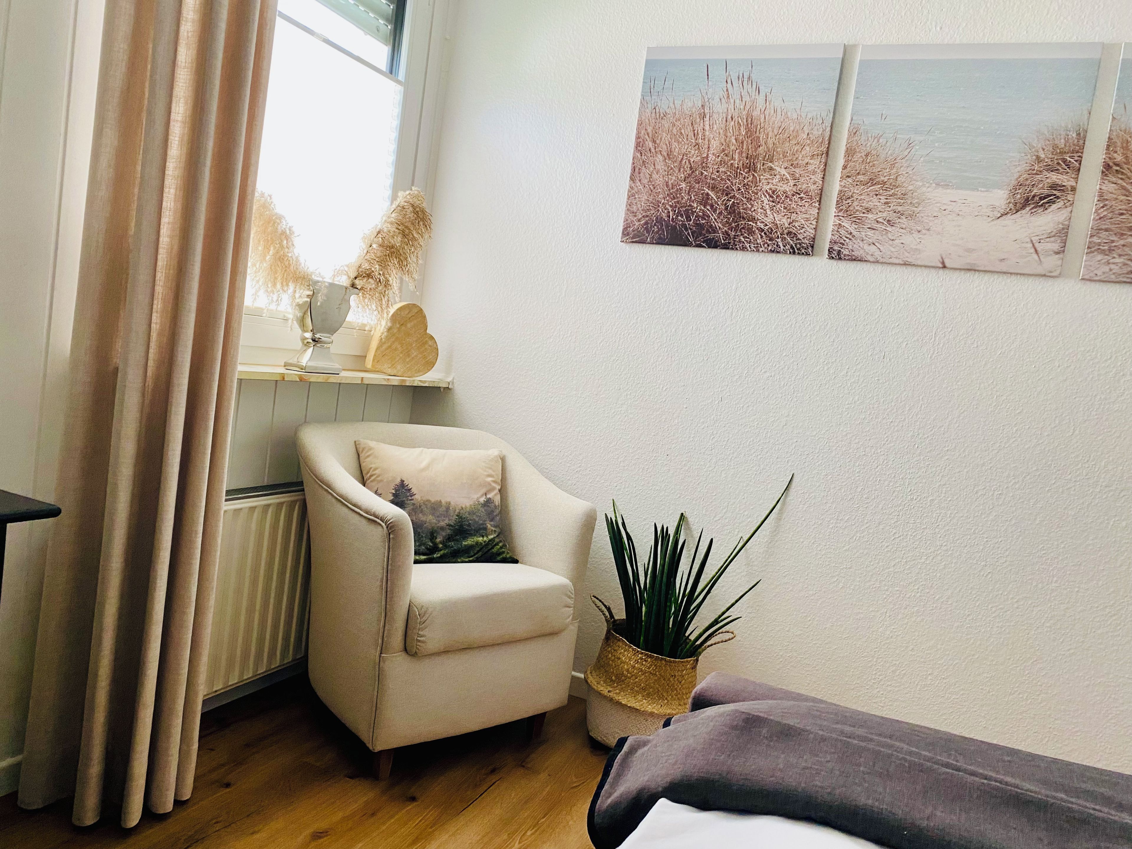 Ein Zimmer mit Holzboden, einem beigefarbenen Sessel mit einem Kissen mit Waldmotiv, einer kleinen Topfpflanze und einer Wand mit drei gerahmten Bildern, die Strandansichten zeigen.