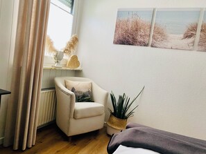 A room with a wooden floor, a beige armchair with a forest-themed pillow, a small potted plant, and a wall with three framed pictures depicting beach scenes.