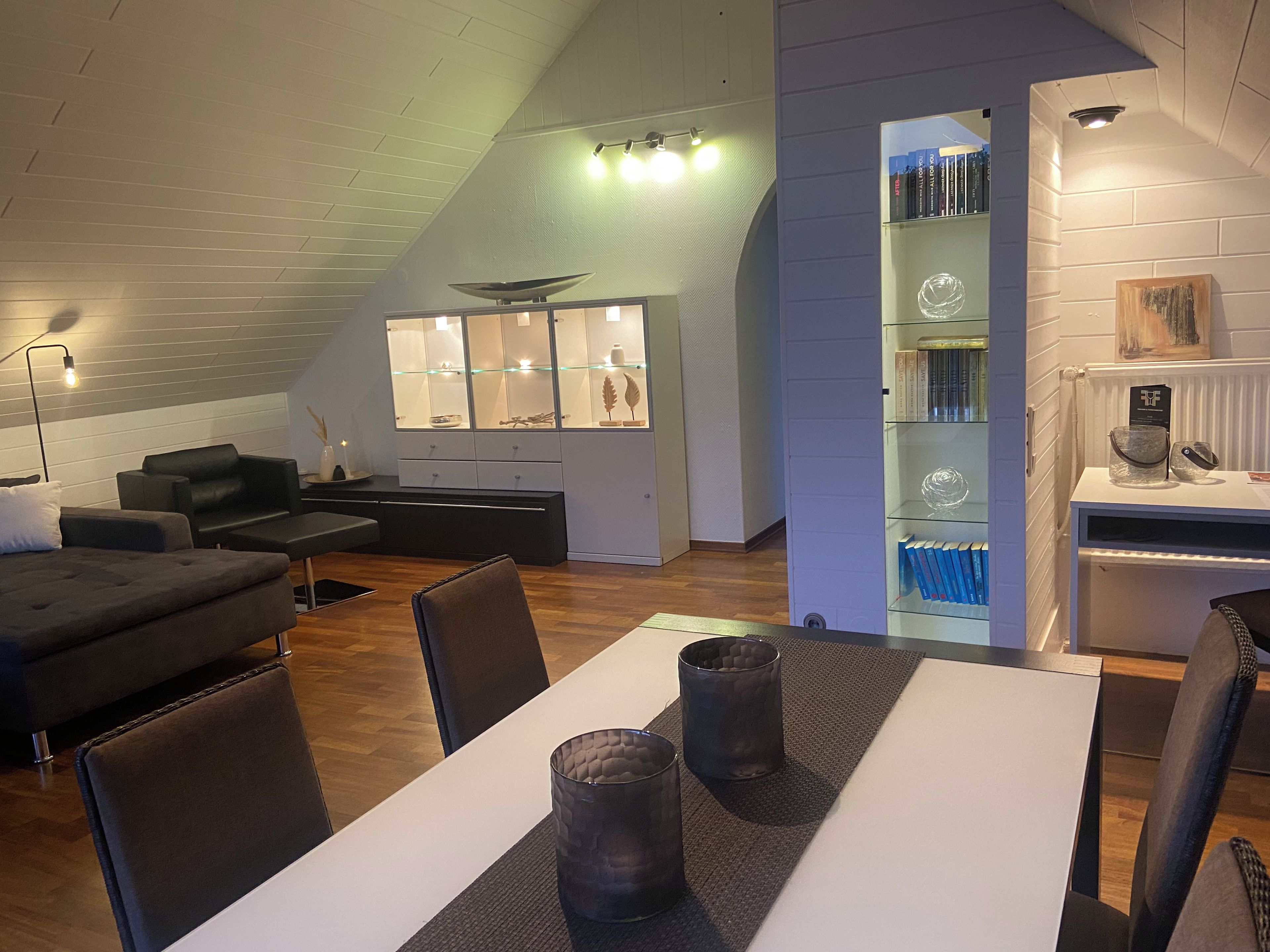 Ein modernes Wohnzimmer mit Essbereich, ausgestattet mit einer Couch, einem Esstisch mit Stühlen, einem Bücherregal und einer dekorativen Glasvitrine.