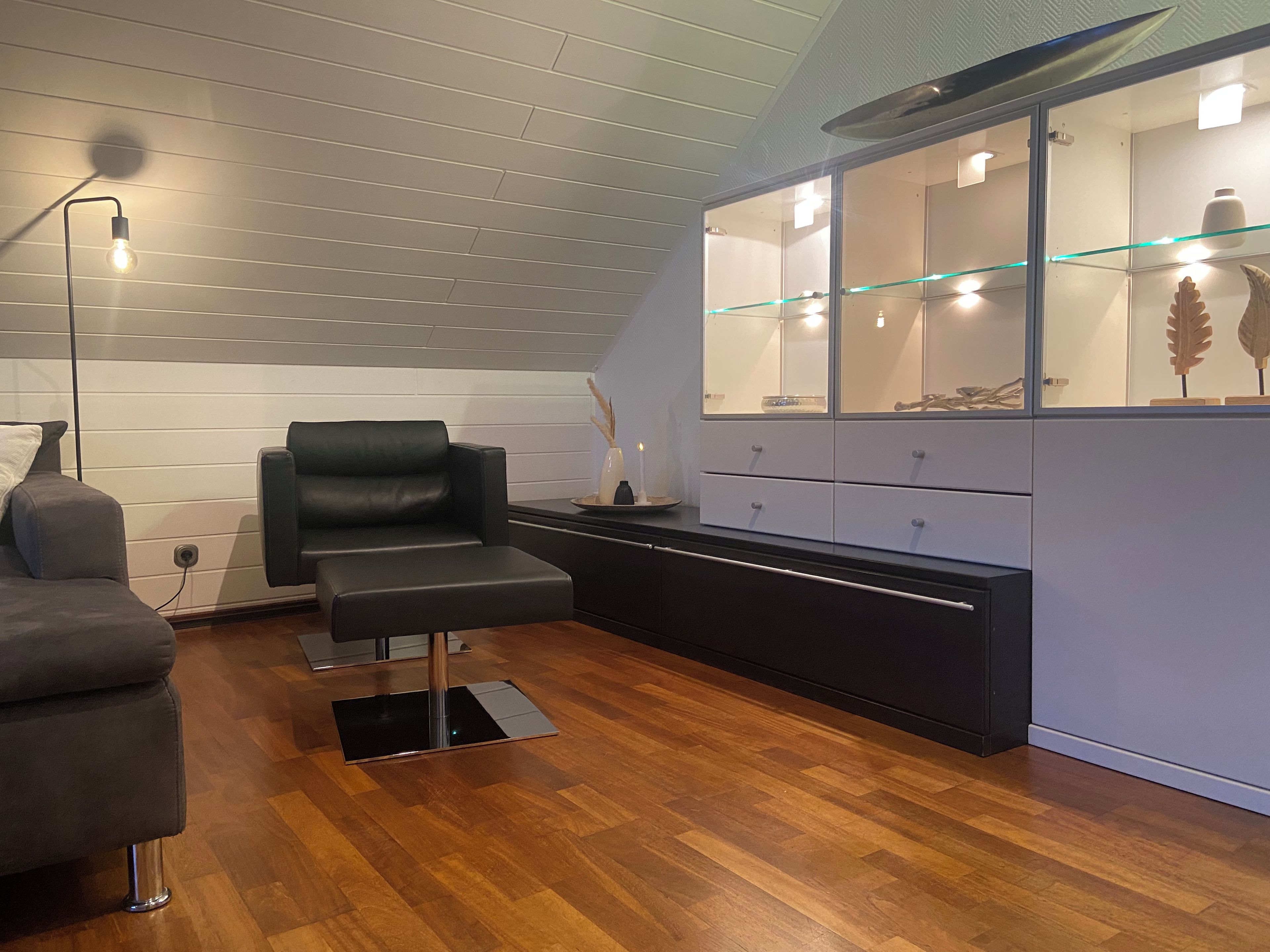 A modern room with a wooden floor, a black leather sofa, a glass cabinet with decorative items, and a standing lamp.