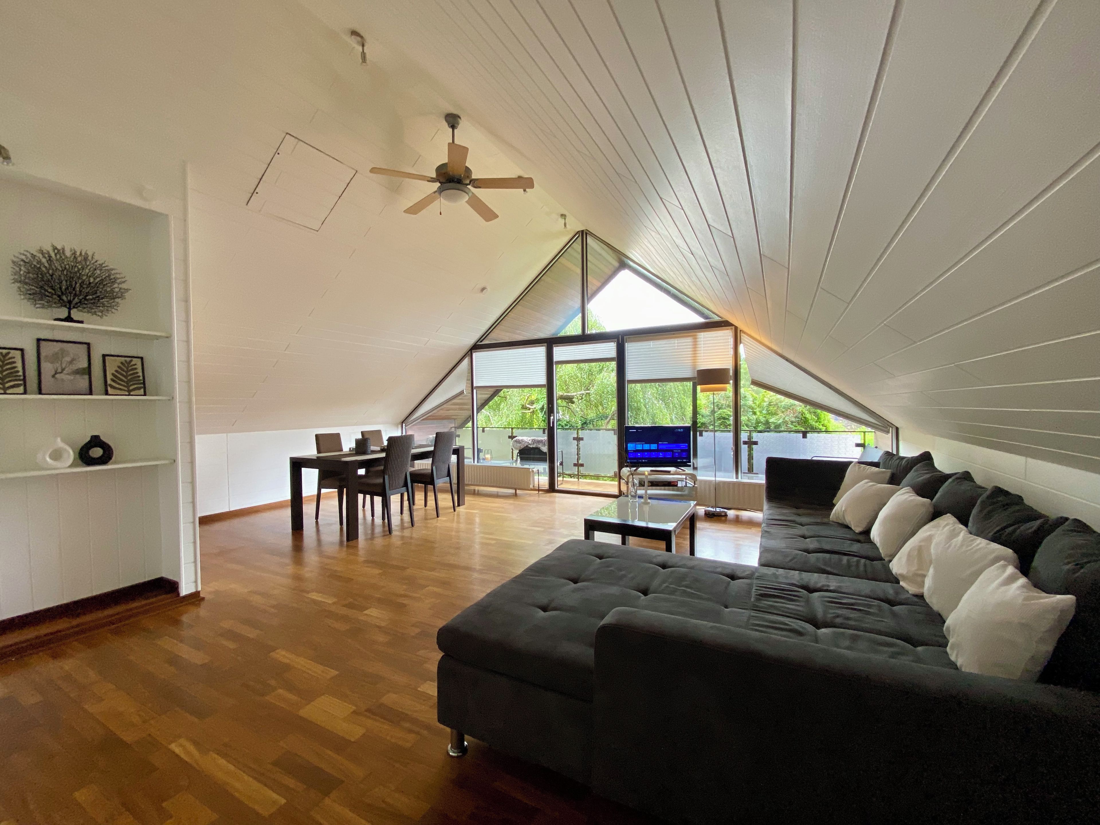 Ein Dachzimmer mit einer dunklen Couch, einem Esstisch und Stühlen. Es gibt einen Deckenventilator, einen Fernseher und ein Fenster mit Blick auf Grünflächen.