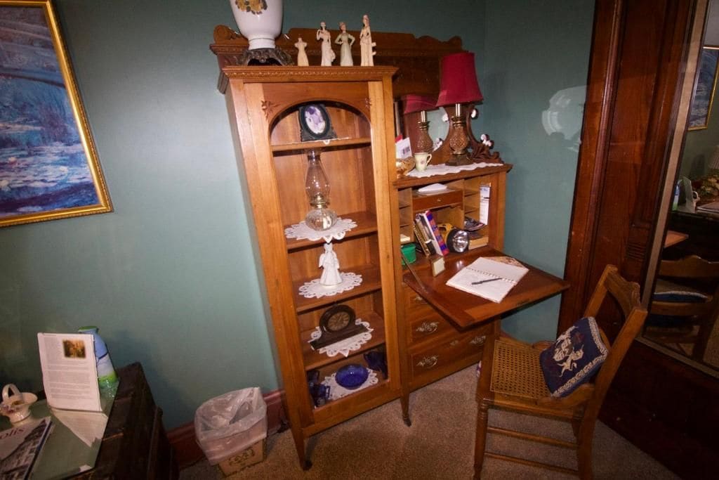 A room with a wooden cabinet, a chair, a table with a lamp, and a painting on the wall.