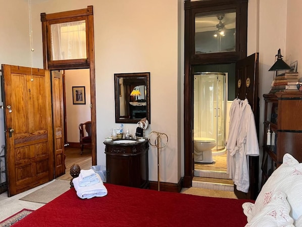 Habitación de hotel con alfombra roja, puerta de madera, baño con cabina de ducha de vidrio y cama con ropa de cama blanca.