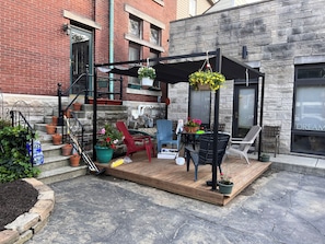 Una terraza de madera con mobiliario exterior, plantas en macetas y un edificio de ladrillo al fondo.