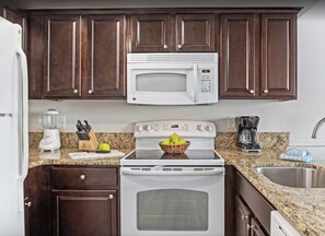 A kitchen with dark wood cabinets, a white microwave, a stove, a sink, and a countertop with a blender and a basket of apples.