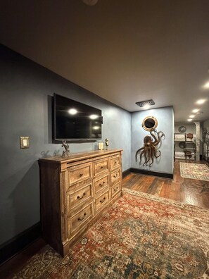 A room with a large flat-screen TV mounted on the wall, a wooden dresser with multiple drawers, a patterned rug on the floor, and a wall-mounted octopus decoration.
