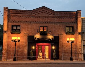 A brick building with a red entrance, two windows with plants, and a number '307' on the door.
