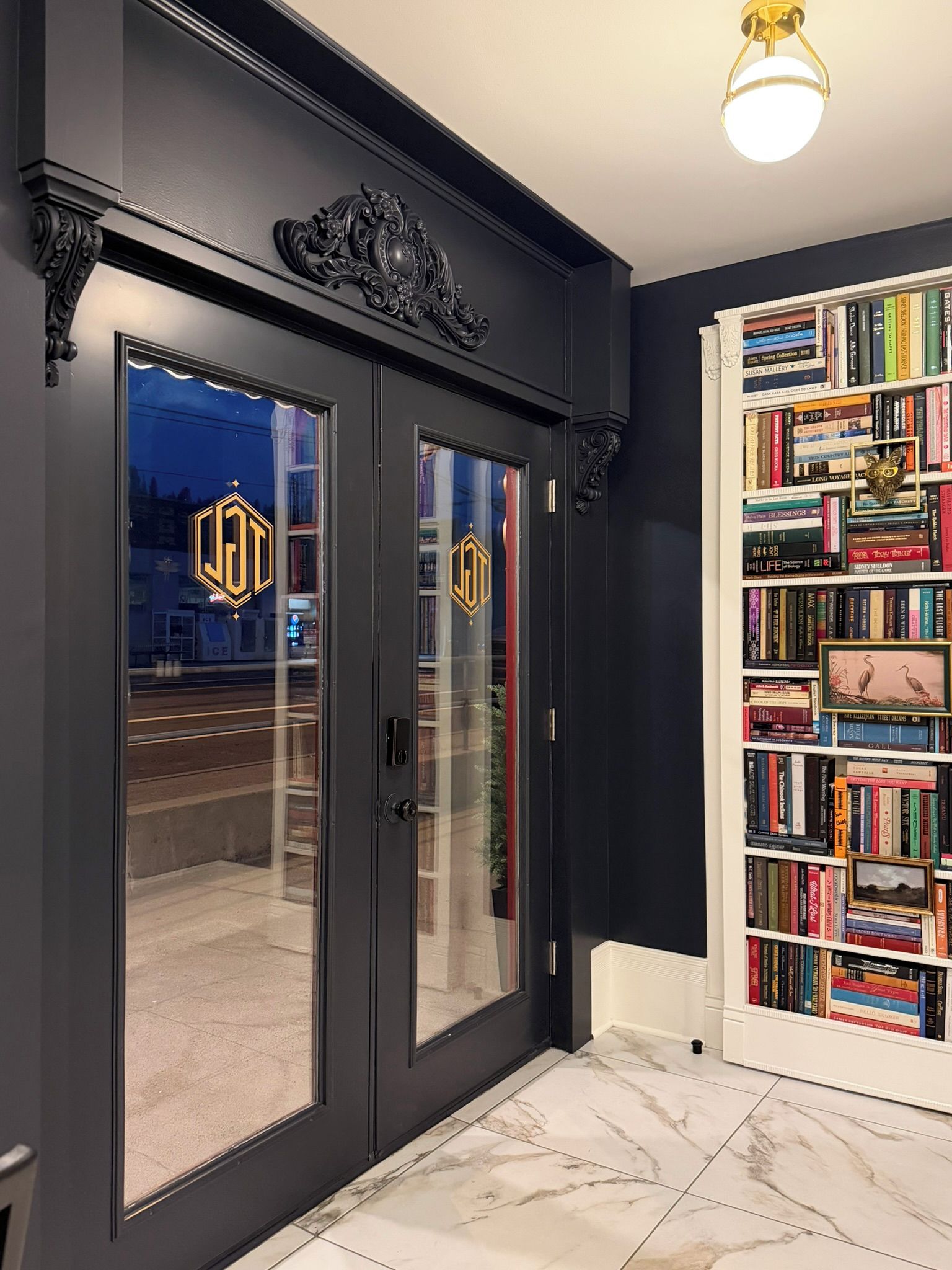 Une porte de couleur sombre, dotée de panneaux de verre et d’un logo, un sol recouvert de carreaux de marbre et une bibliothèque remplie de livres.