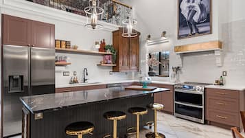 Een moderne keuken met een groot eiland, barstoelen, roestvrijstalen apparatuur en een decoratieve plafondlamp.