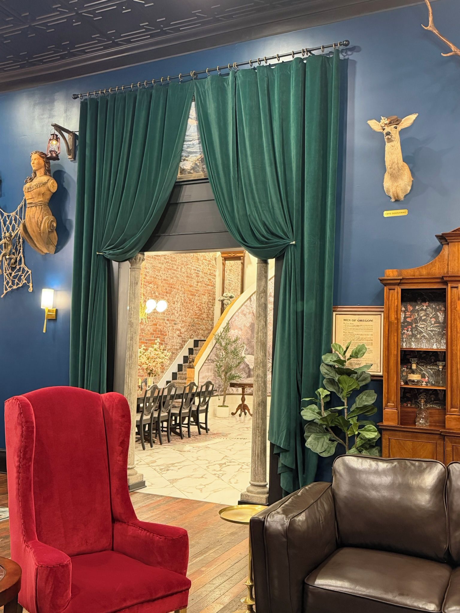 Een kamer met een rode stoel, een bruine leren bank, een groen gordijn, een aan de muur bevestigd hertshoofd en een trap naar een buitenruimte.