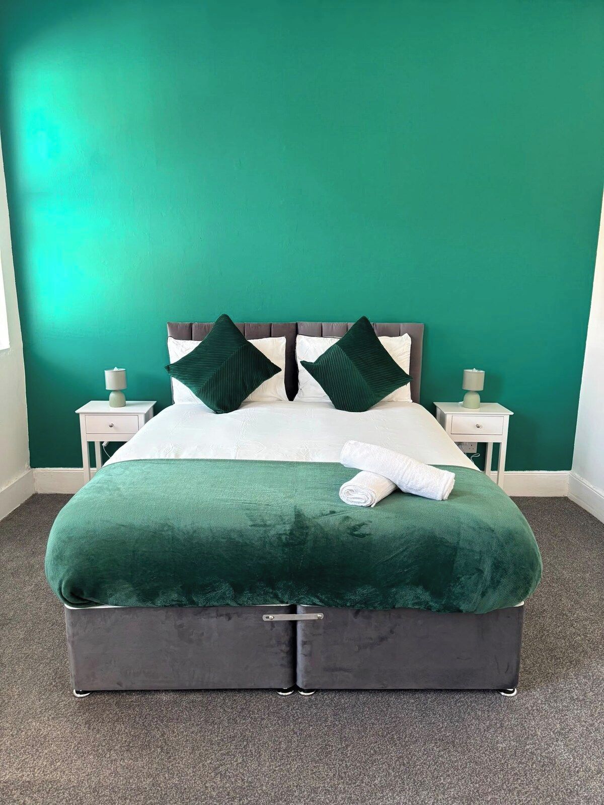 Ein Bett mit grünem, bezogenen Kopfteil, weißen Bettwäsche und einer grünen Tagesdecke. Es befinden sich zwei Nachttische mit Lampen davor.