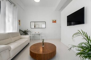 Ein modernes Wohnzimmer mit einem weißen Sofa, einem runden Holztisch, einem kleinen Tisch mit Stühlen, einem flachbildfernseher an der Wand und einer Topfpflanze.