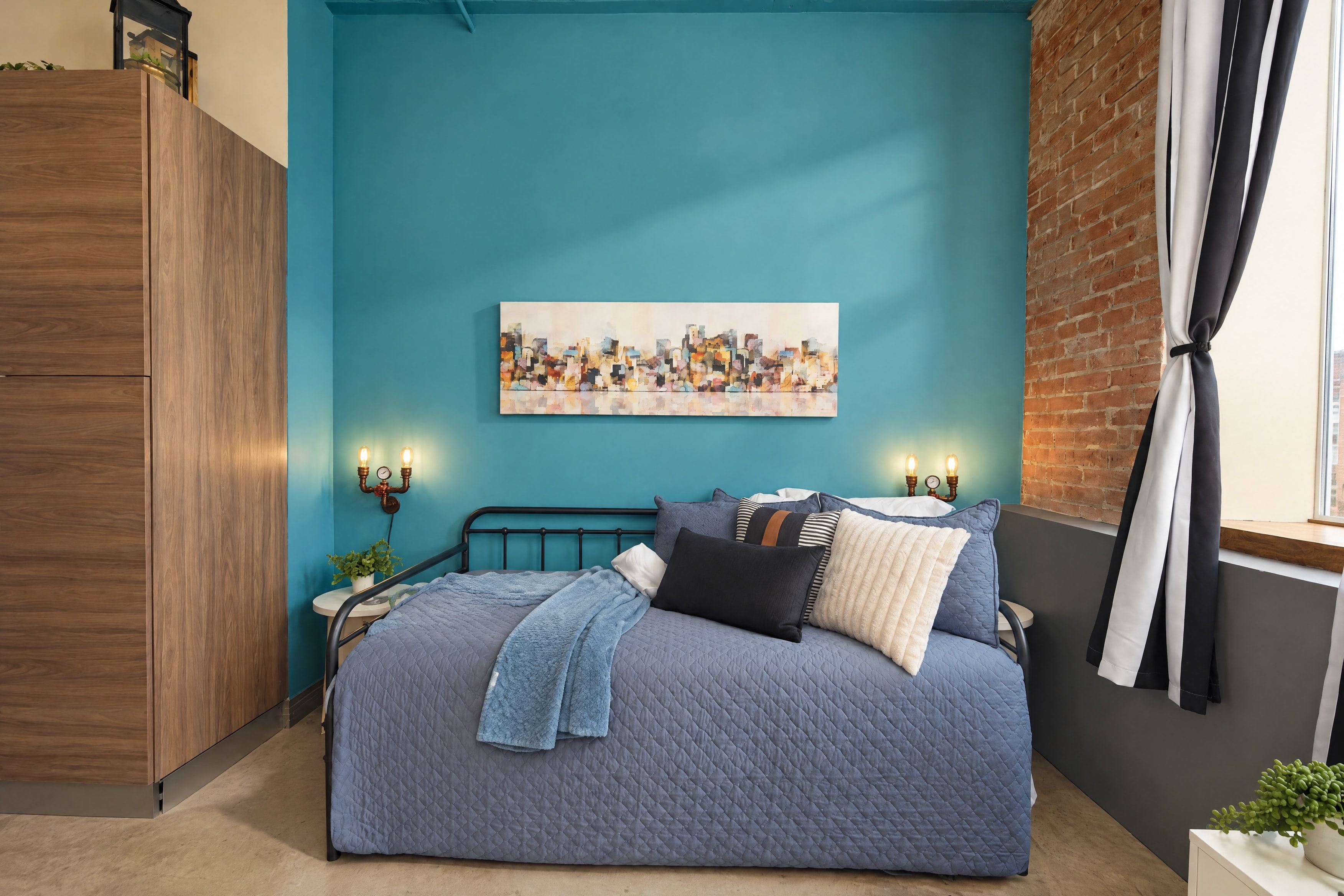 A bedroom with a bed, a cityscape painting, bedside lamps, and a brick wall.