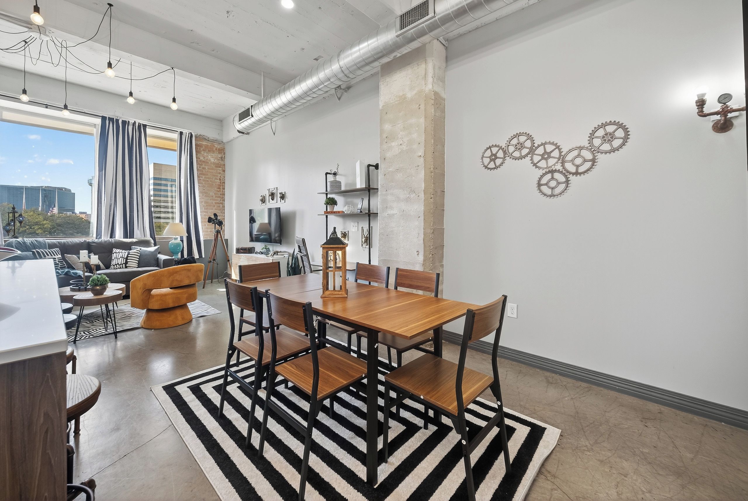 A modern living room with a dining area, featuring a large wooden table, black and white striped rug, and a city view through large windows.