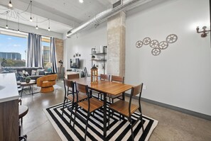 A modern living room with a dining area, featuring a large wooden table, black and white striped rug, and a city view through large windows. - The Bentley Collection - The Meridian (Dallas)