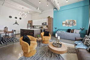 A modern living room with a sofa, armchairs, and a coffee table. There is a kitchen area with cabinets and a dining table. - The Bentley Collection - The Meridian (Dallas)