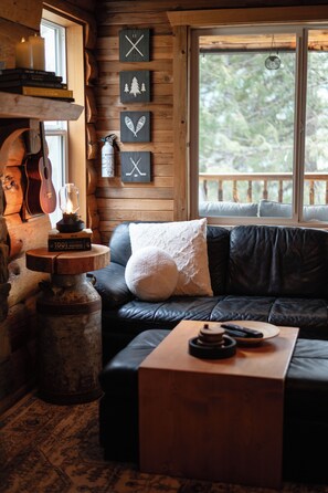 A cozy cabin interior with a wooden wall, a black leather sofa, a small wooden table, and a window with a view of trees.