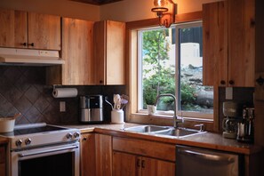 A kitchen with wooden cabinets, a stainless steel coffee maker, and a window with a view of greenery. - Snow & Soak | Private Hot Tub, Sauna & Cold Plunge Near Manning Park (Manning Park)