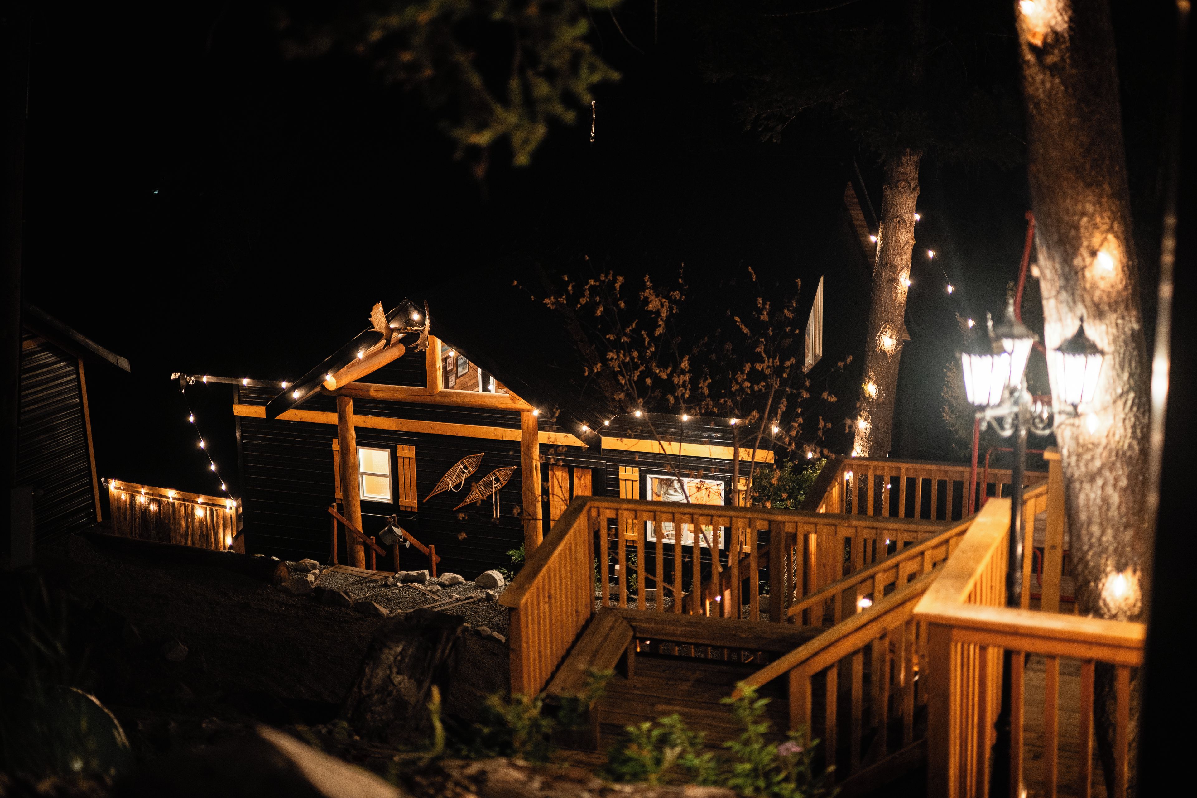 Une cabane en bois avec un escalier menant à une terrasse, entourée d’arbres et illuminée de guirlandes lumineuses.