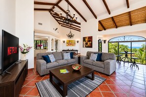 A spacious living room with a large flat-screen TV, two sofas with patterned cushions, a coffee table, and a view of lush greenery through a glass door. - Ocean View Villa • Tamarindo 5 min • Walk to Beach (Tamarindo)