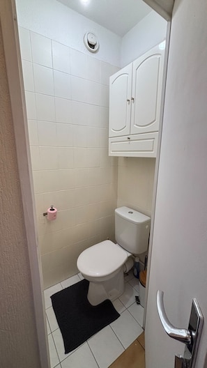 Ein kleines Badezimmer mit Toilette, einem weißen Schrank darüber und einer schwarzen Matte auf dem Boden.