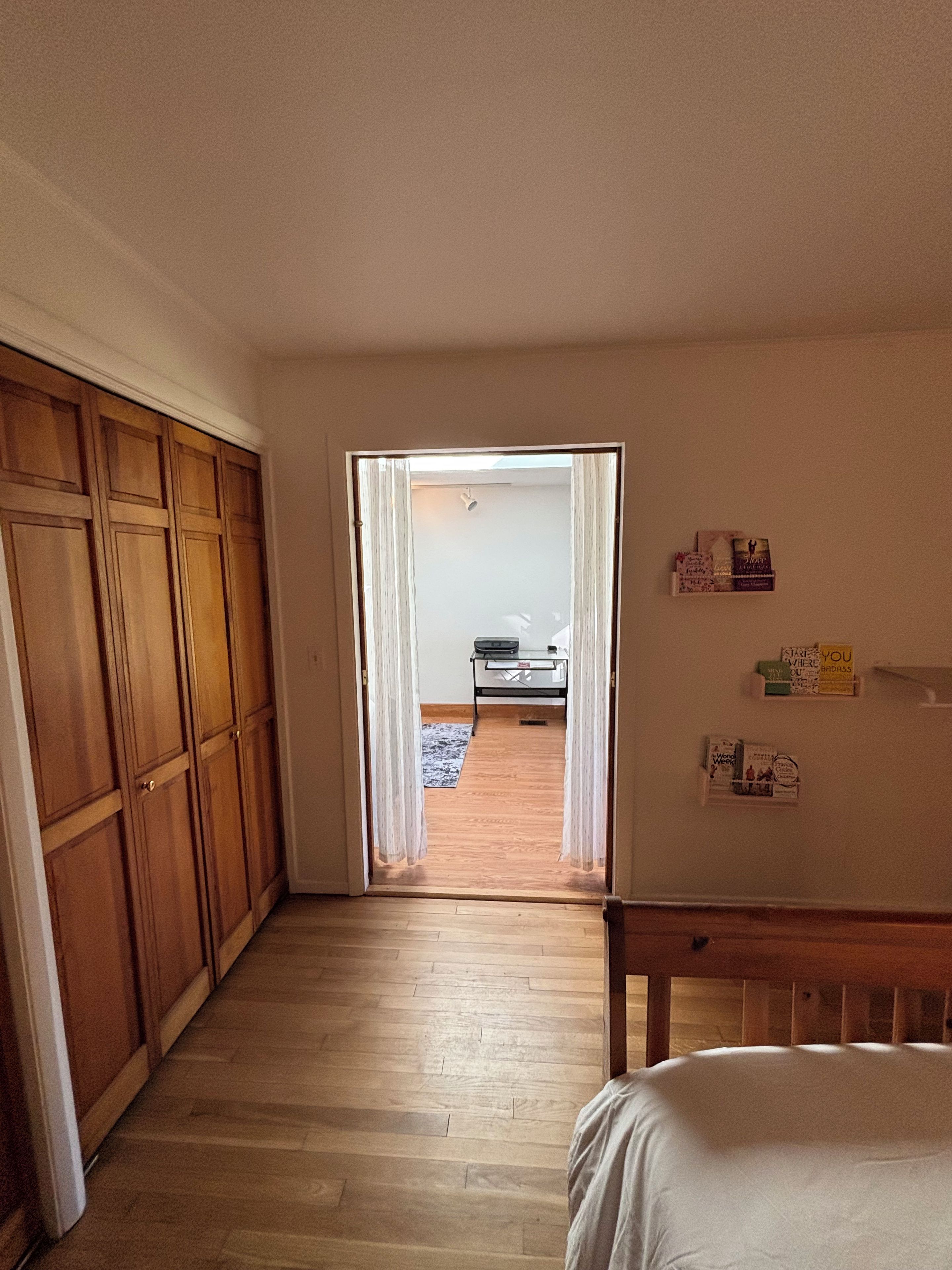 Un couloir mène à une chambre qui contient une armoire en bois, un lit et une petite table avec une lampe.
