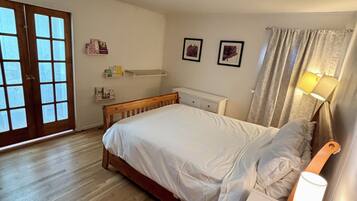 Een slaapkamer met een houten bed, een nachtkastje met een lamp, een raam met gordijnen en een deur met glaspanelen.