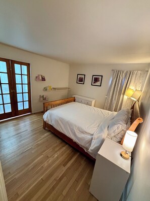 Un dormitorio con cama de madera, una mesita de noche con lámpara, un ventanal con cortinas y una puerta con paneles de vidrio.