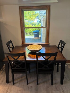 Une table à manger agrémentée d’un élément décoratif tissé, entourée de quatre chaises, placée devant une fenêtre donnant sur une poubelle bleue et de la verdure à l’extérieur.