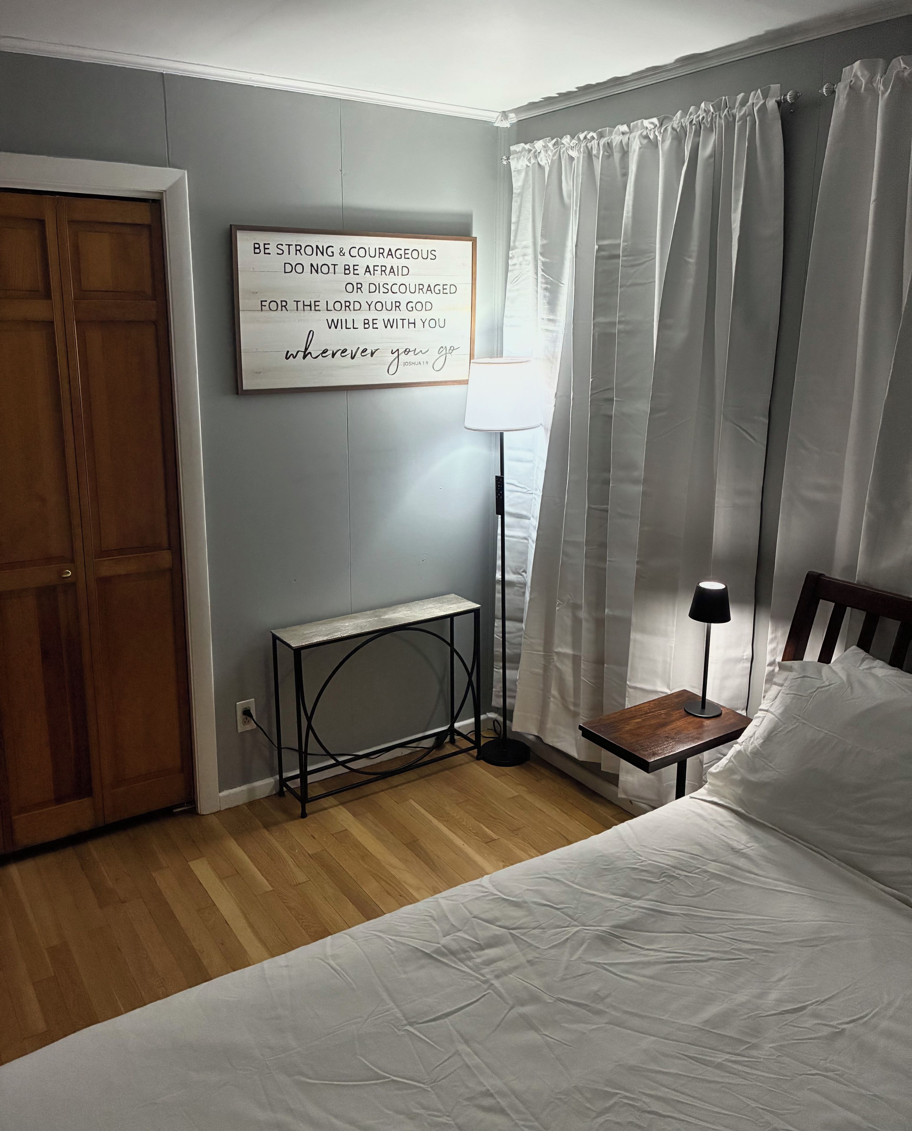 Une chambre à coucher avec un lit, une table de chevet, une lampe, un panneau fixé au mur et une porte en bois.
